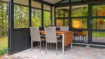 A covered patio with a table and chairs, large windows, and a view of greenery.