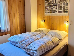 A room with wooden panel walls, a bed with a floral duvet, two bedside lamps, and a poster of townhouses. - 4 Person Holiday Home in Farvang (Farvang)