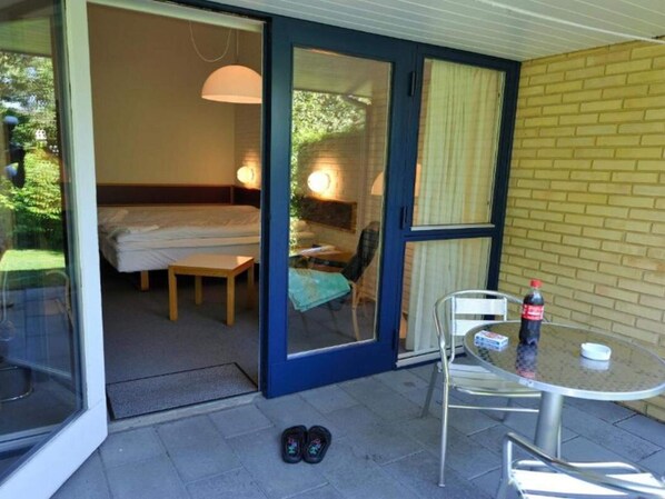 A room with a bed, a small table, and a chair. There is a glass door leading to an outdoor area with a table and a chair.