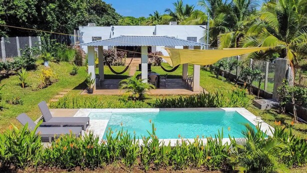 Un espace piscine doté d’un coin salon abrité, entouré d’une végétation luxuriante et avec un bâtiment blanc en arrière-plan.