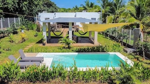 A pool area with a covered seating area, surrounded by lush greenery and a white building in the background. - The Pacific in Salinitas (Acajutla)