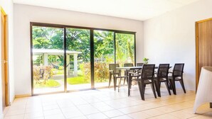 Une pièce moderne dotée d’une grande porte-fenêtre coulissante donnant sur un jardin, d’une table à manger avec des chaises et d’un sol carrelé de couleur blanche.