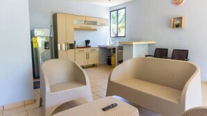 Un espace de vie compact comprenant un coin cuisine, deux fauteuils en osier, une petite table et un lit simple.