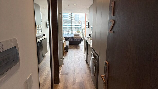 A modern apartment interior with a wooden floor, a kitchen with a refrigerator and oven, a dining area, and a large window offering a city view.