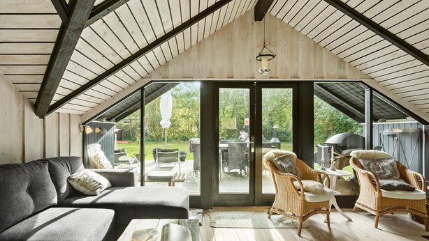 A cozy attic room with a wooden ceiling, a large sliding glass door leading to an outdoor patio, a grey sofa, wicker chairs, and a coffee table with fruits. - Beautiful home in Gedser with WiFi (Gedser)