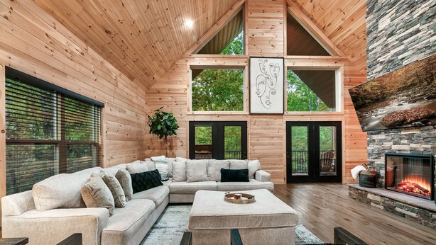 Interior acogedor de una cabaña de madera con un sofá modular, una chimenea y ventanas amplias con vistas a un bosque.