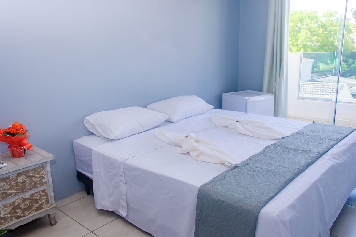 A neatly made bed with white linens and a gray blanket, a small bedside table with a vase of flowers, and a window with a view of greenery outside.
