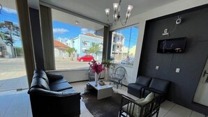 A modern living room with a black leather sofa, a wooden chair, and a coffee table. A TV is mounted on the wall, and there is a large window providing a view of the street outside. - Hotel Caçula Do pampa (Sao Gabriel)