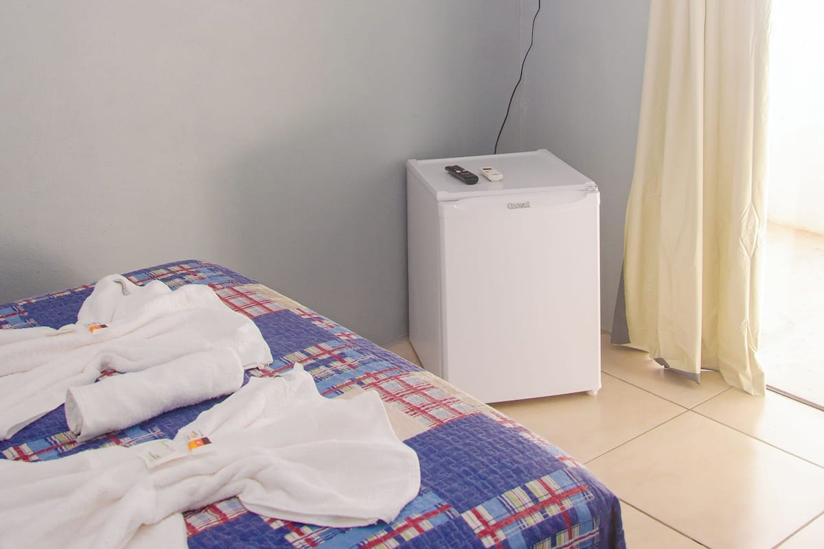A hotel room with a bed, a flat-screen TV mounted on the wall, a white air conditioner, and a window with curtains.