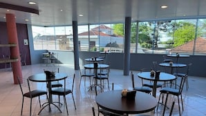 A spacious indoor area with multiple round tables and chairs, large windows offering a view of buildings and trees, and a tiled floor. - Hotel Caçula Do pampa (Sao Gabriel)