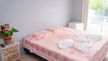 Una cama bien hecha con ropa de cama y toallas con motivos florales, una planta en la mesita de noche y un pequeño aparador blanco con cajones.