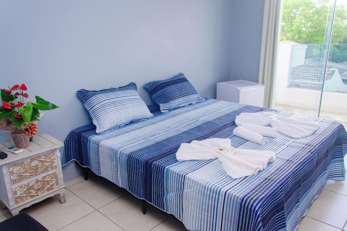 A neatly made bed with blue and white striped bedding, a bedside table with a plant, and a window with a view of buildings.