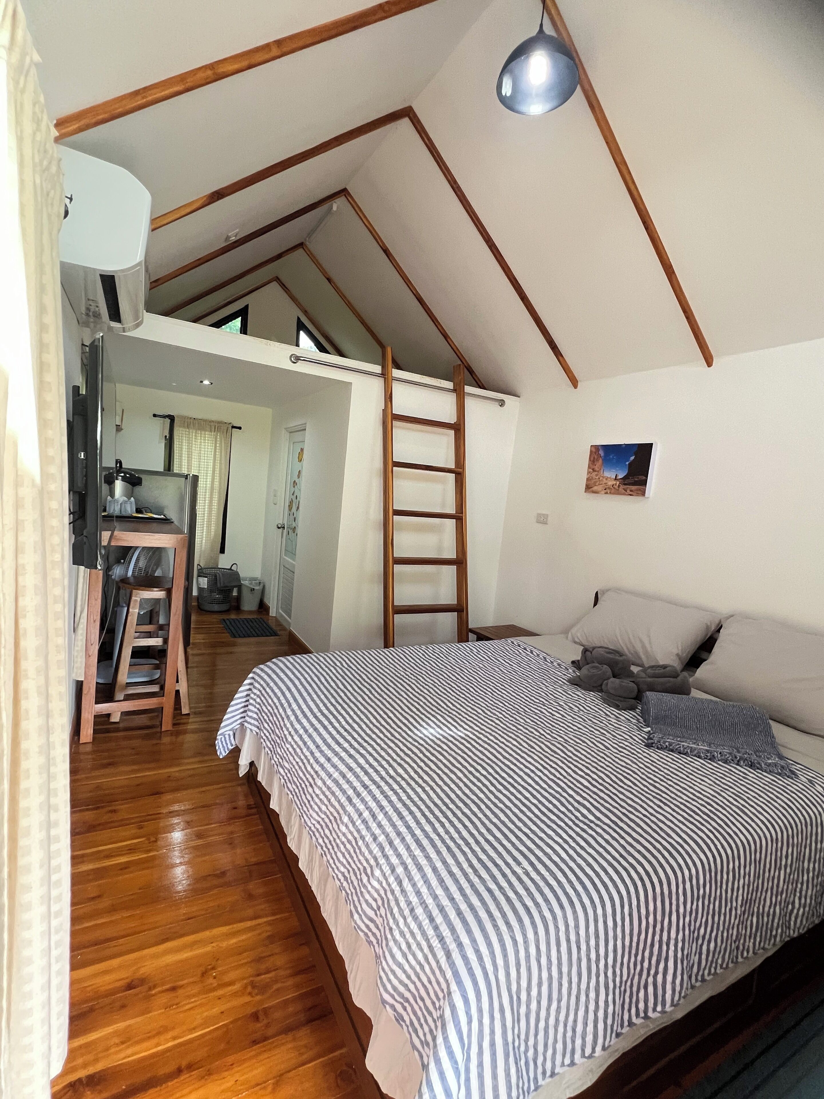 A bedroom with a lofted sleeping area, wooden beams, a bed with striped bedding, a ladder, and a picture on the wall.