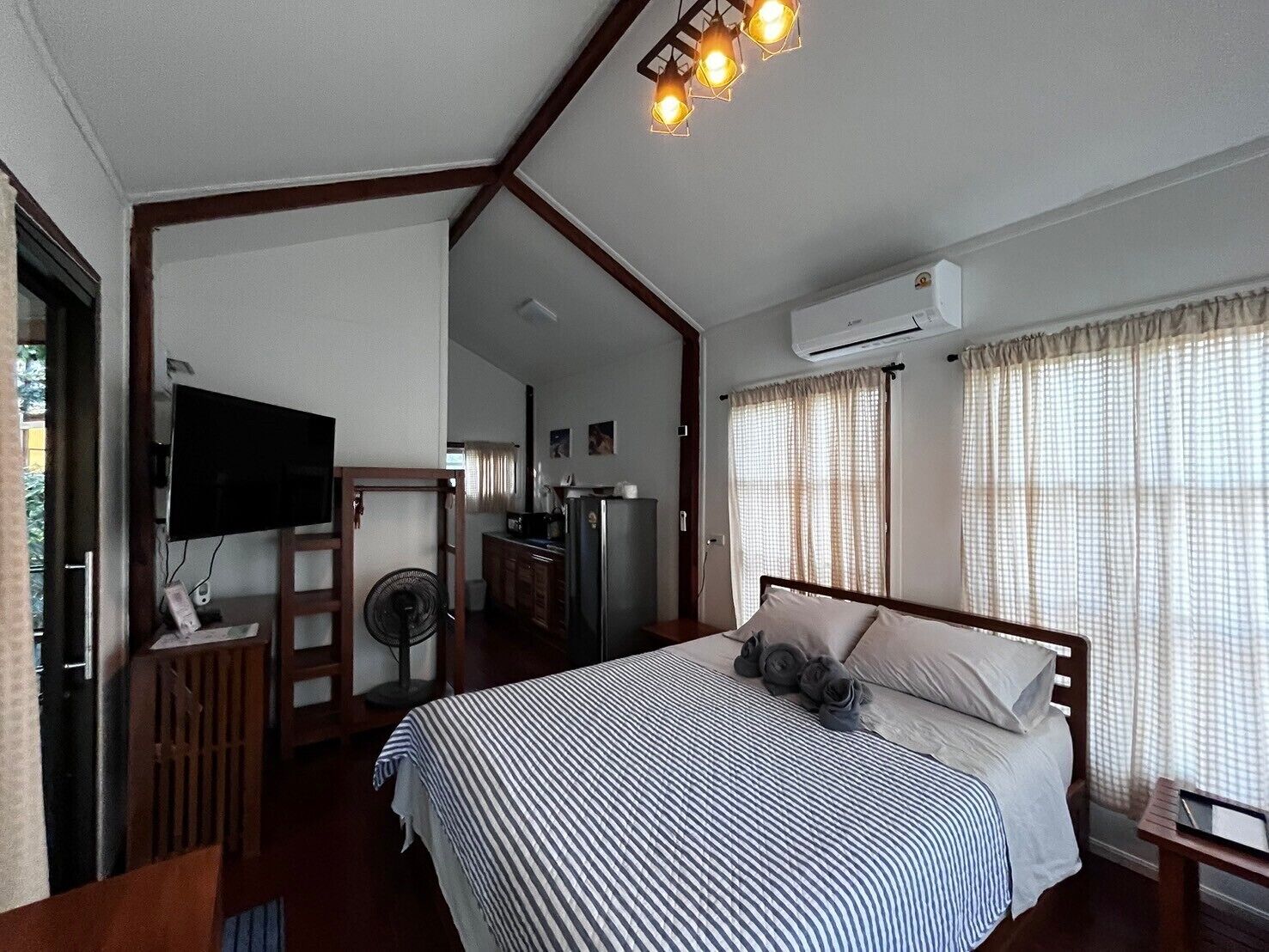 Un dormitorio con cama, televisión, ventilador y una kitchenette al fondo.