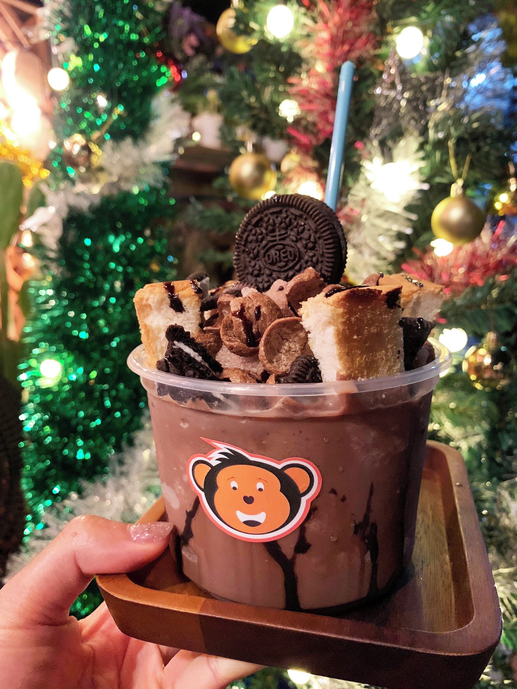 A cup of chocolate drink with cookies and a bear sticker, held by a hand in front of a Christmas tree.