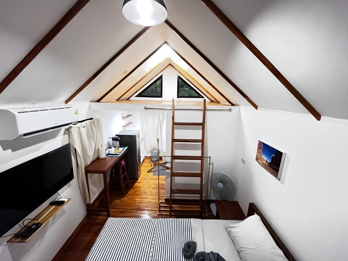 Ein gemütlicher Raum mit schrägen Wänden, einem Etagenbett, einem Schreibtisch mit Stuhl, einer Leiter, einem Ventilator und einem Fenster.