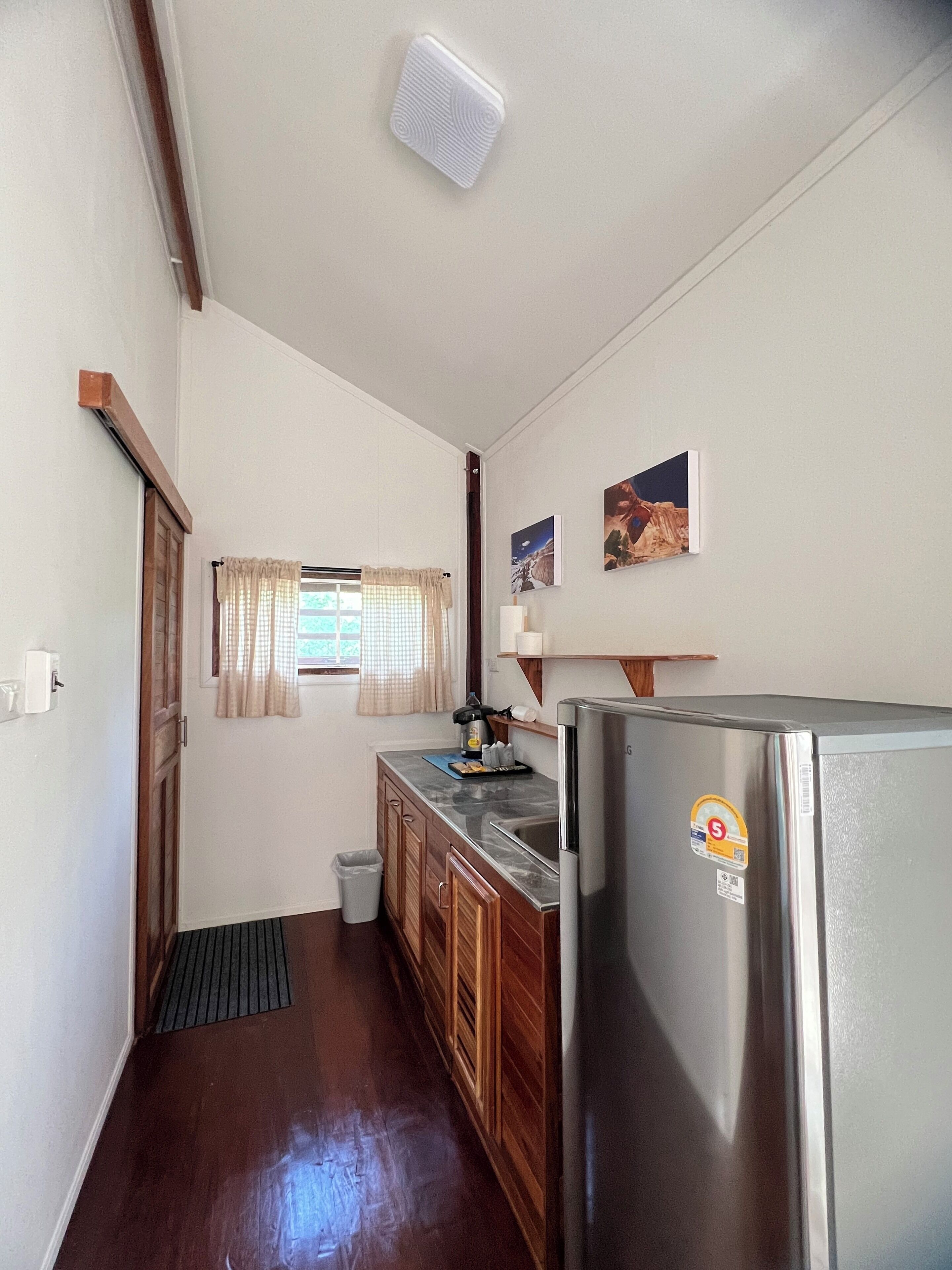 Cozinha compacta com armários de madeira, geladeira de aço inoxidável, janela com cortinas e uma pequena lixeira.