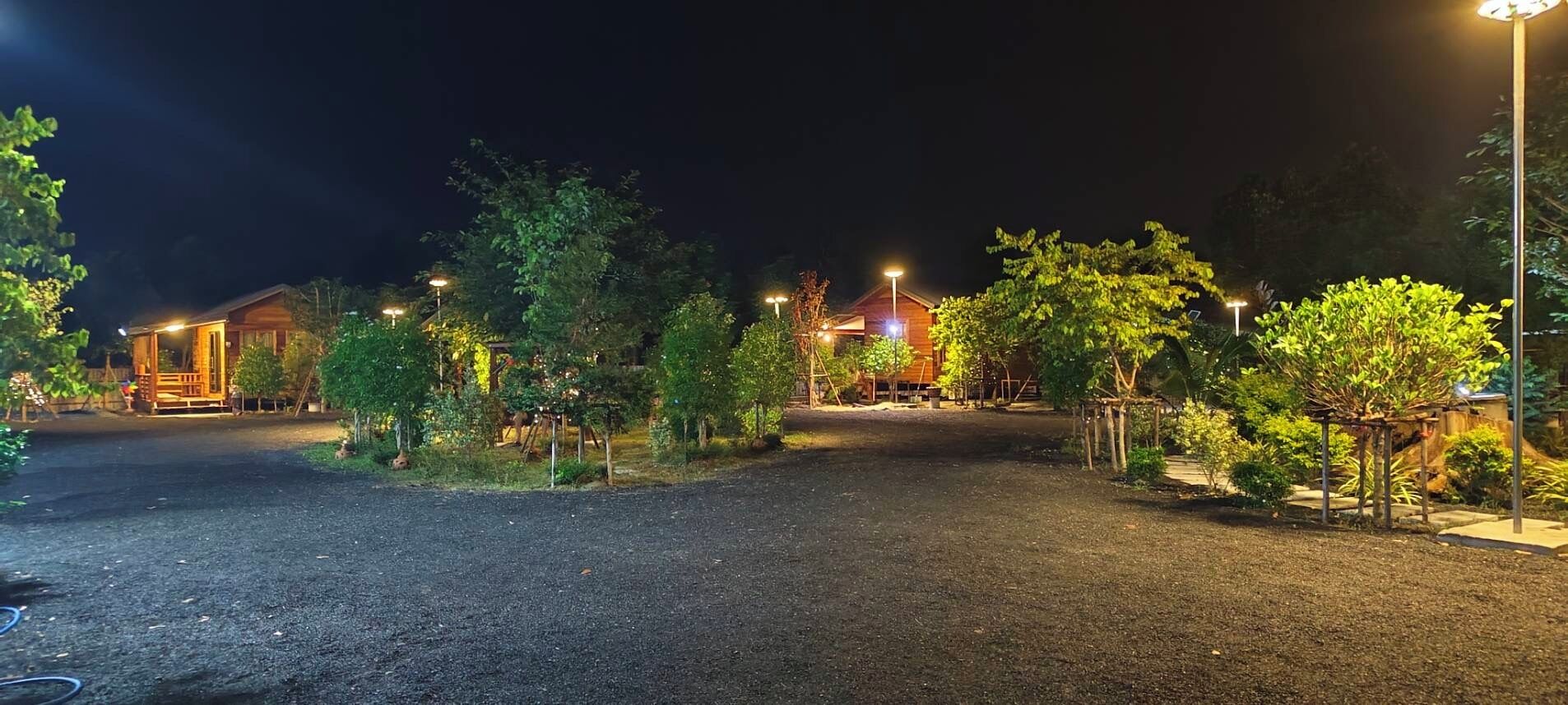 A nighttime view of a well-lit outdoor area with a gravel path, surrounded by trees and wooden structures.