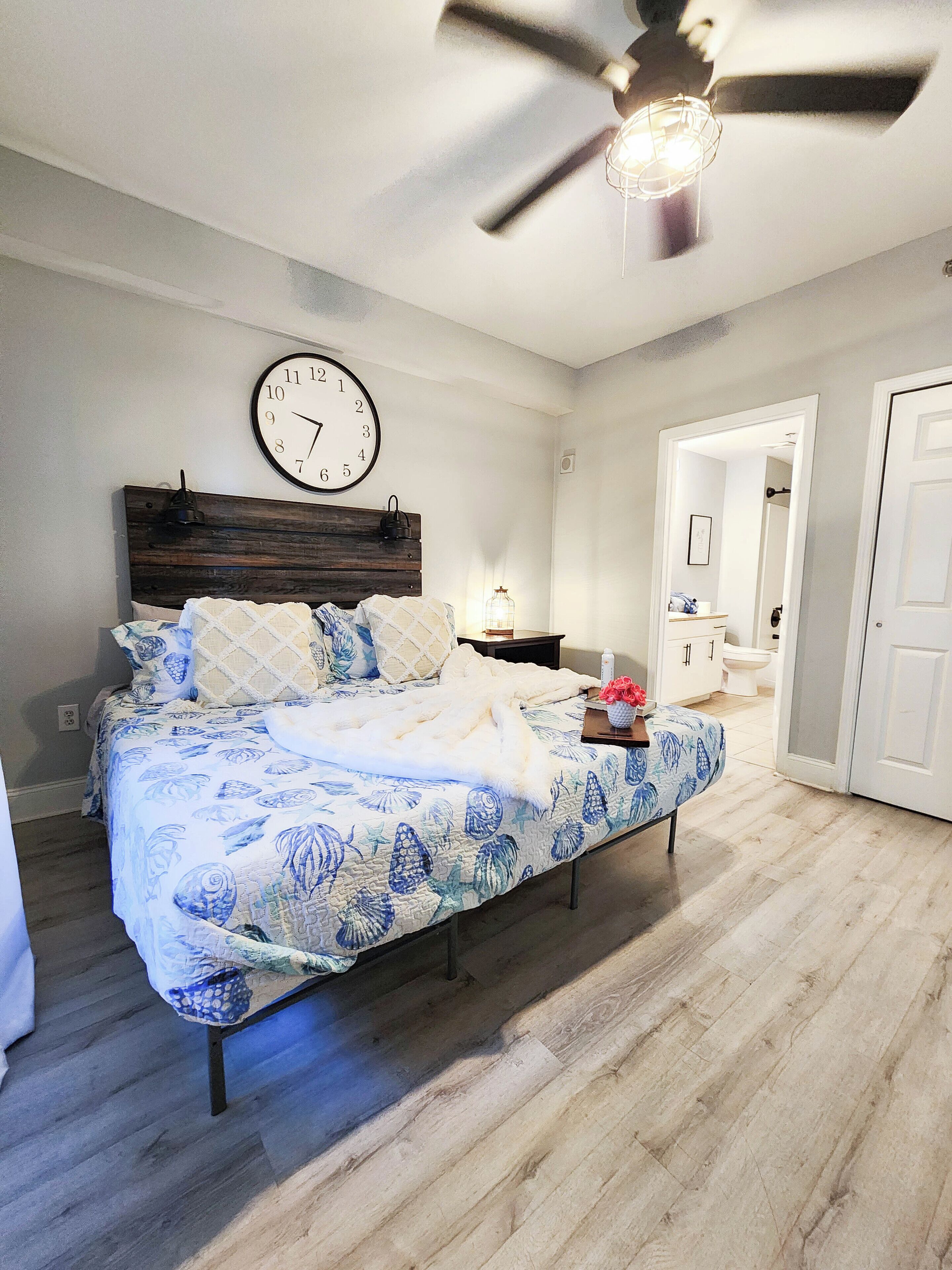 A bedroom with a bed, a wall clock, a ceiling fan, and a nightstand with a lamp.