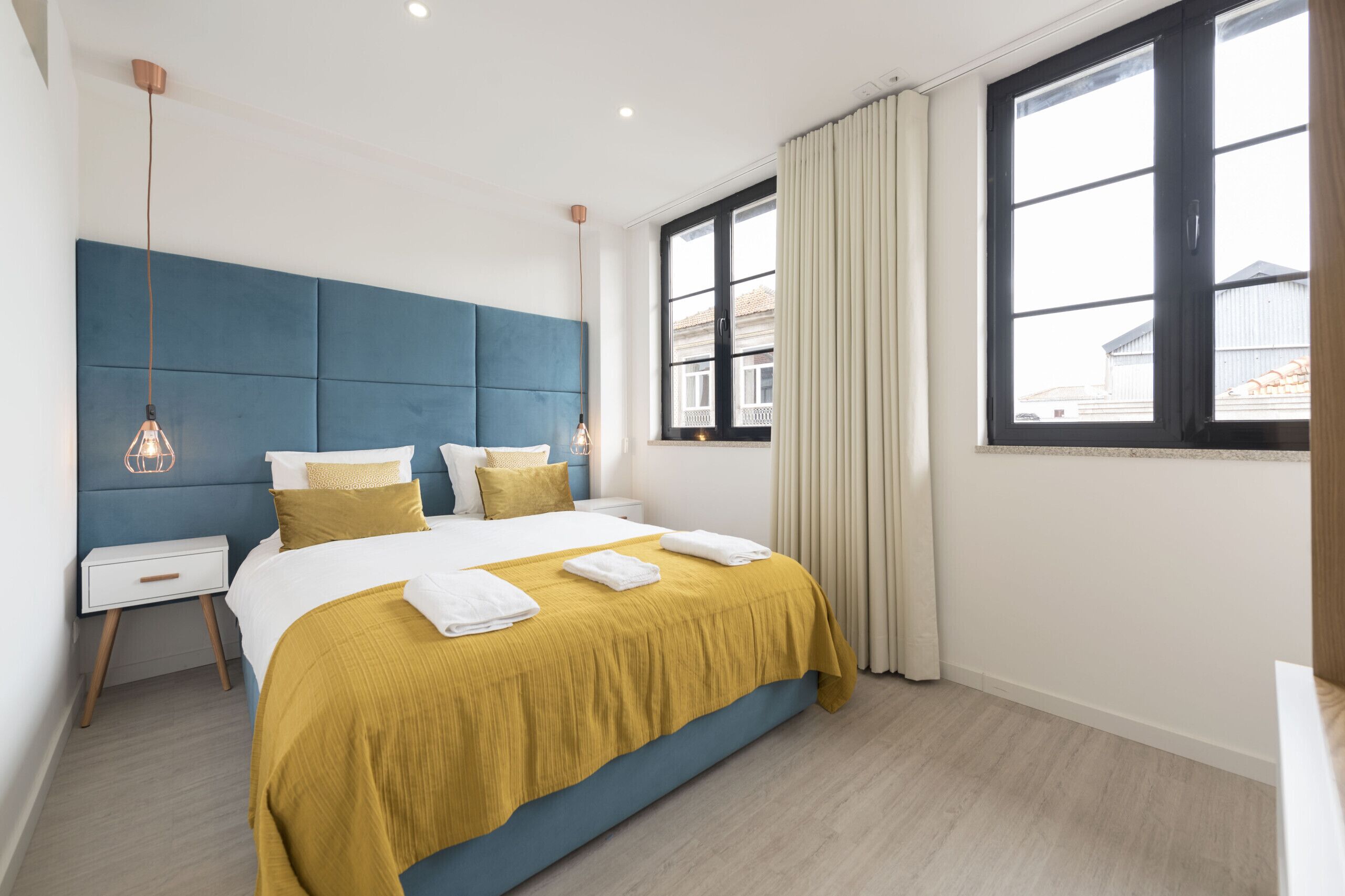 Ein modernes Schlafzimmer mit einem großen Bett, einem blauen Kopfteil, einem Nachttisch und einem Fenster mit Vorhängen.