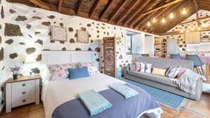 A bedroom with a stone wall, wooden beams, a bed, a sofa, and a kitchenette. - Holiday Home 'Estudio Bibi' with Mountain Views and Private Terrace (El Paso)