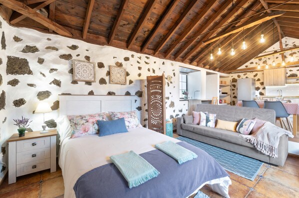 A bedroom with a stone wall, wooden beams, a bed, a sofa, and a kitchenette.