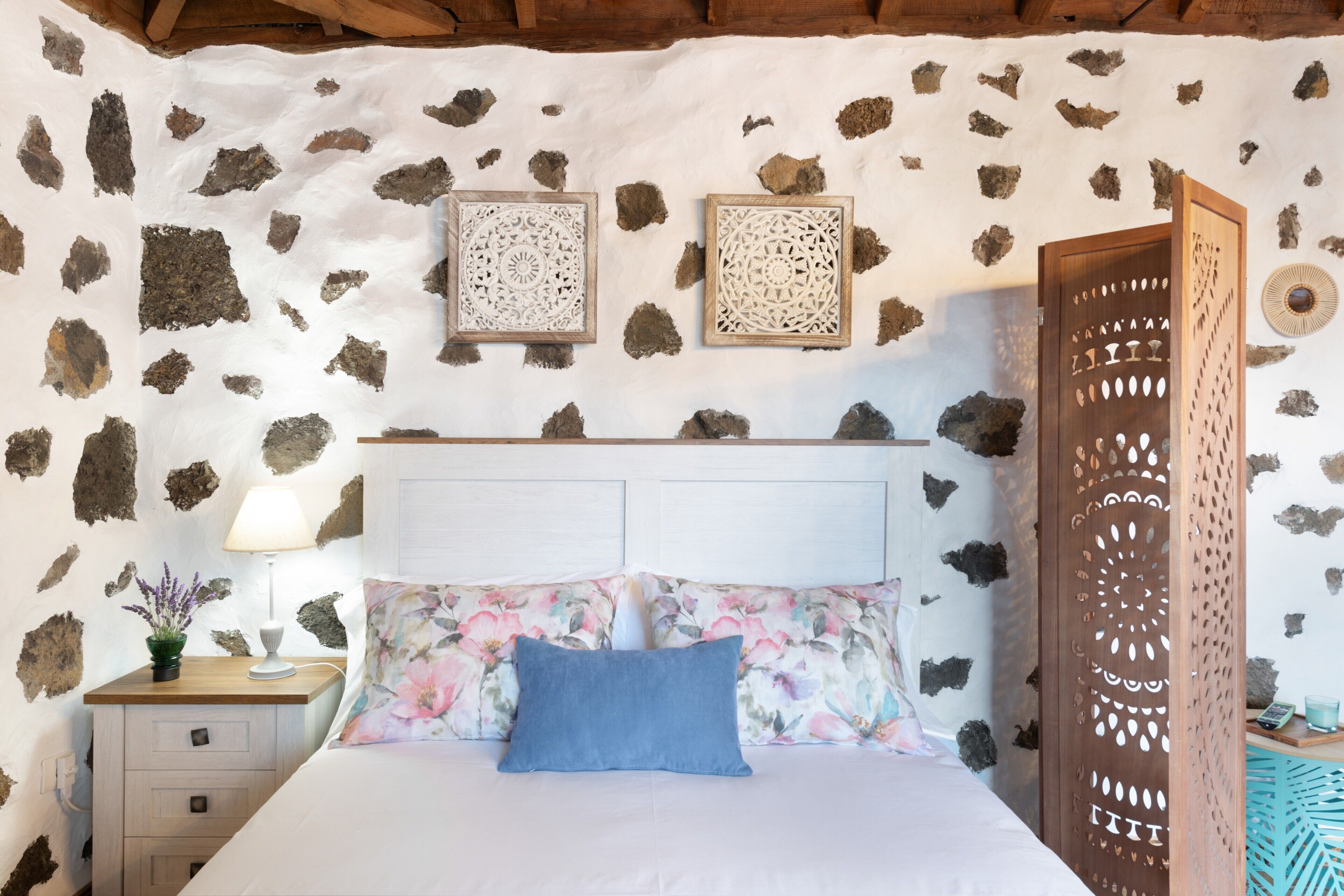 Un dormitorio con pared de piedra, cabecera de madera, colcha estampada con motivos florales y mesita de noche con lámpara.