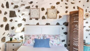Un dormitorio con pared de piedra, cabecera de madera, colcha estampada con motivos florales y mesita de noche con lámpara.