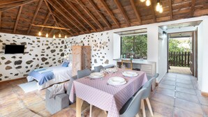 A rustic room with a stone wall, wooden ceiling beams, a bed, a dining table set for four, and a view through a door to an outdoor area. - Holiday Home 'Estudio Bibi' with Mountain Views and Private Terrace (El Paso)