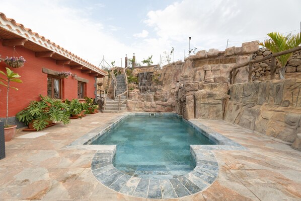 A swimming pool with a stone wall, potted plants, and a red building with a tiled roof.