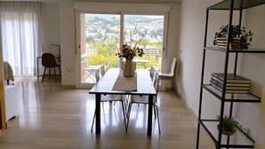 A modern living room with a dining area, a balcony with a city view, and a bookshelf with books and plants.