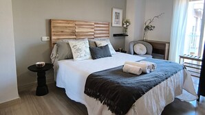 A bedroom with a wooden headboard, a bed with white and dark bedding, a bedside table, a small round table, and a hanging light fixture.