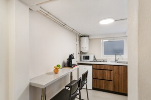 A compact kitchen with a microwave, sink, and a small refrigerator. There is a window above the sink and a countertop with chairs.