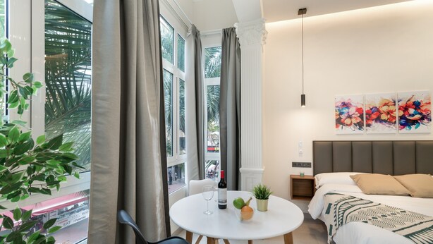 A modern hotel room with a bed, a round dining table with chairs, a potted plant, a wine bottle, and a painting on the wall. - Vacation Studio 'Estudio Esquina' with Shared Terrace, Wi-Fi and Air Conditioning (Las Palmas de Gran Canaria)