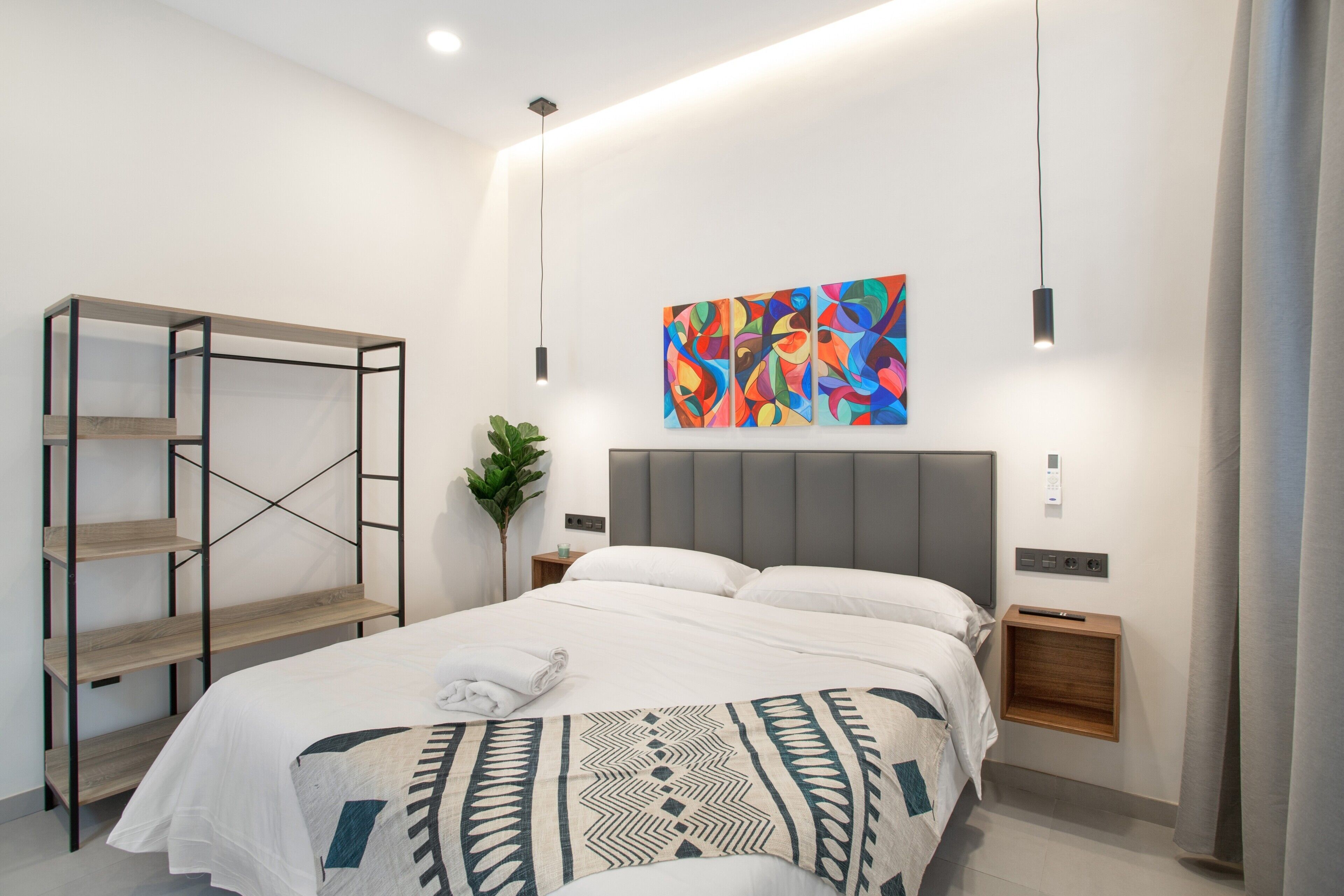 A modern hotel room with a bed, a wall-mounted shelf, a colorful abstract painting, and a bedside table with a plant.