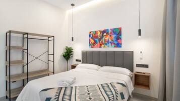 A modern hotel room with a bed, a wall-mounted shelf, a colorful abstract painting, and a bedside table with a plant.