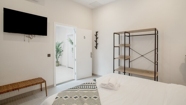 Ein Schlafzimmer mit Bett, einem an der Wand montierten Fernseher, einer Holzbank, einer Pflanze und einer Tür zu einem anderen Raum.