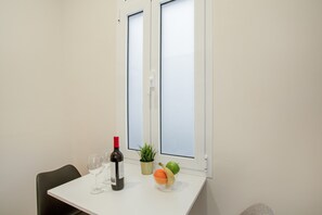 A room with a white table, a bottle of wine, a bowl of fruit, a potted plant, and a chair.