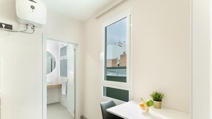 Una habitación moderna con un escritorio blanco, una silla, una planta en maceta y una ventana con vistas a edificios.
