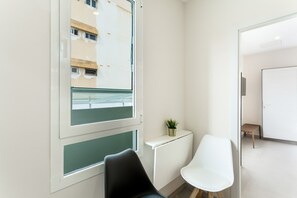 Een moderne kamer met een groot raam, een witte stoel, een klein tafeltje met een plant en een witte kast.