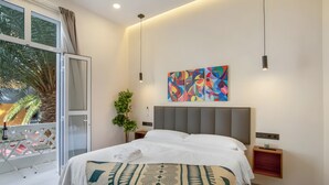 A modern hotel room with a large bed, a balcony with a view of palm trees, and a colorful abstract painting on the wall.