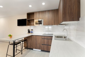A modern kitchen with wooden cabinets, a built-in microwave, a stainless steel sink, and a flat-screen TV mounted on the wall.