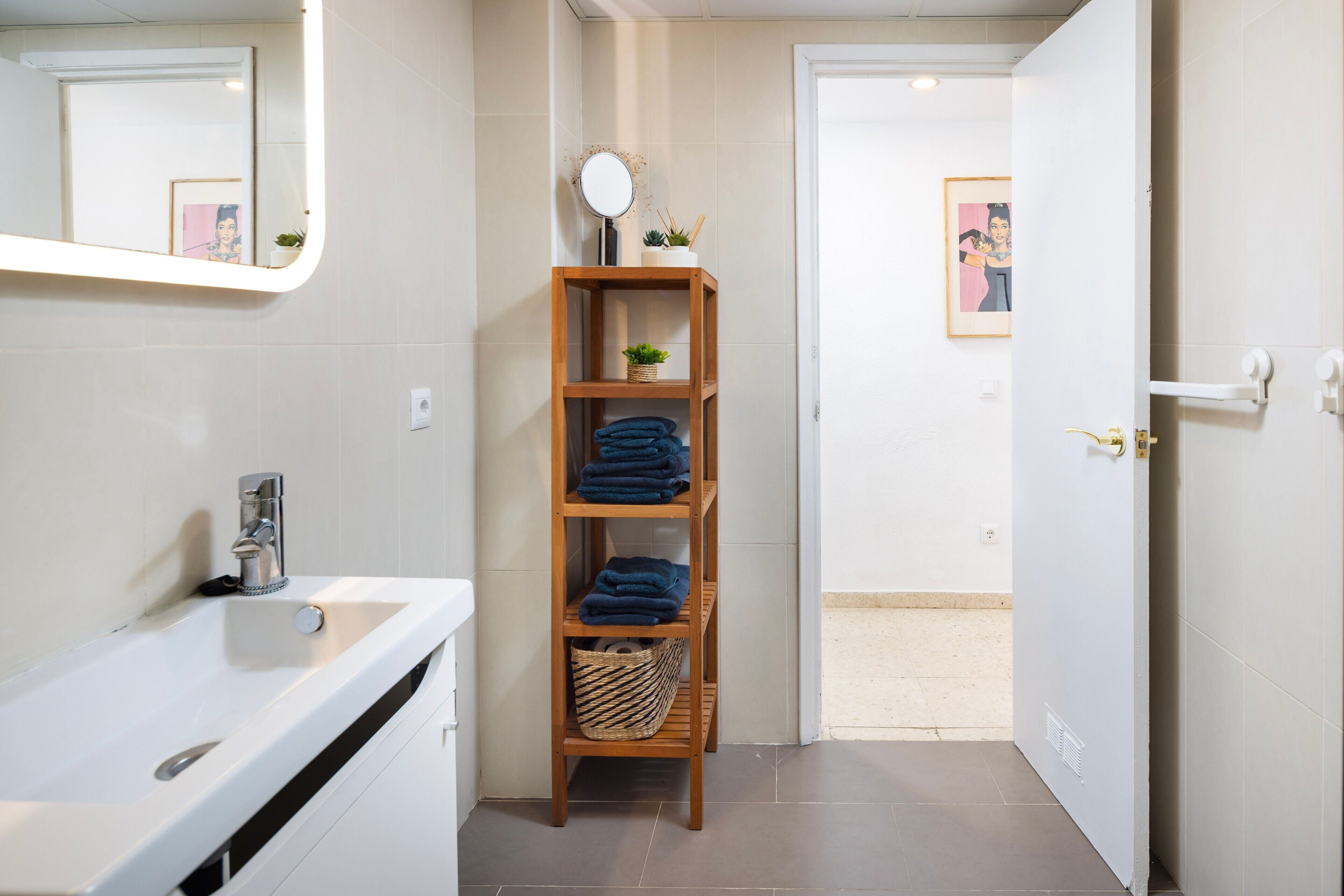 Une salle de bain avec un lavabo blanc, une étagère en bois où sont pliées des serviettes, un petit miroir et un tableau encadré accroché au mur.