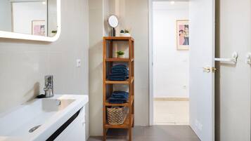 Une salle de bain avec un lavabo blanc, une étagère en bois où sont pliées des serviettes, un petit miroir et un tableau encadré accroché au mur.