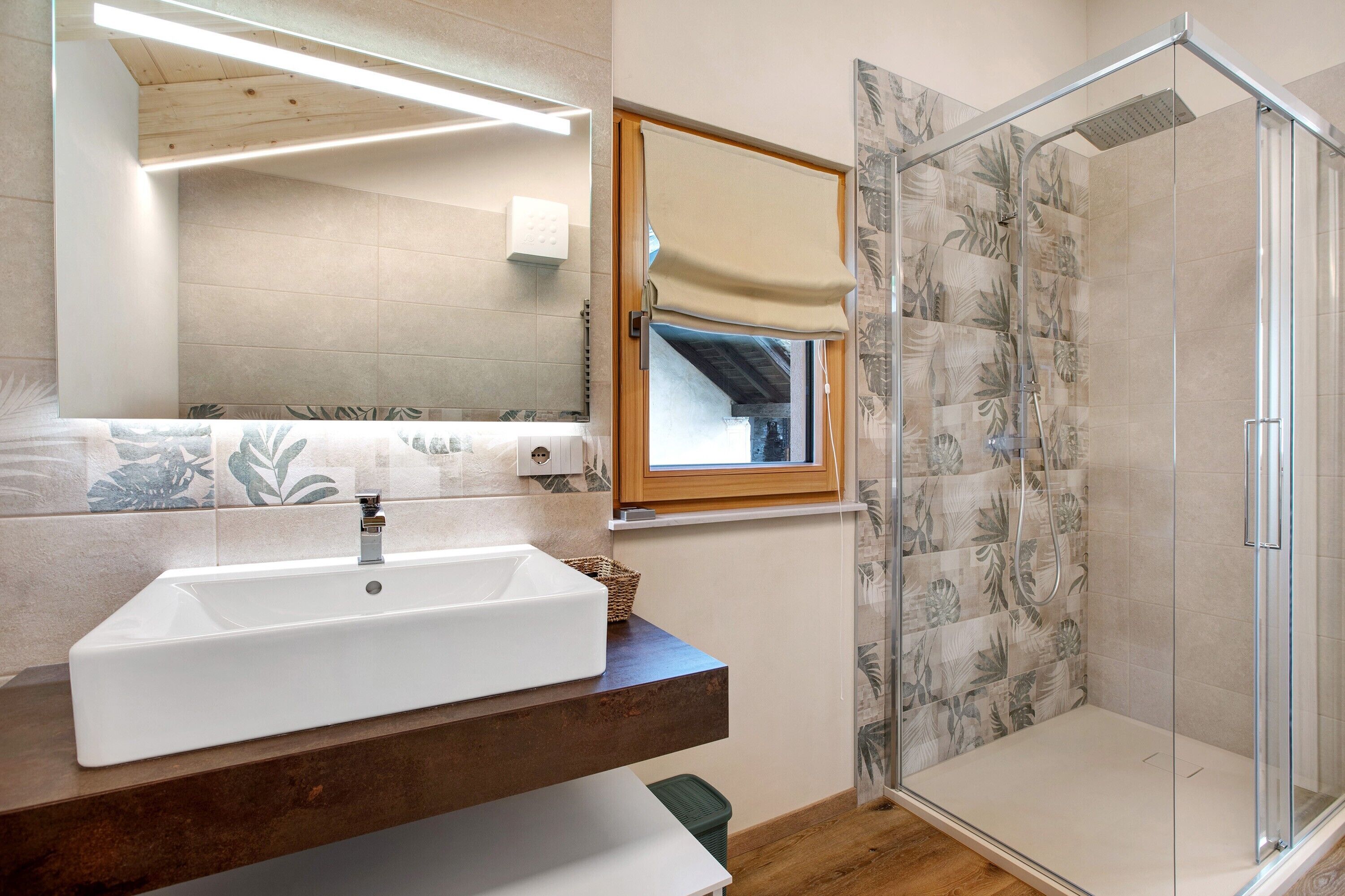 Une salle de bain avec un lavabo blanc, un miroir, une douche avec une porte en verre et une fenêtre avec un cadre en bois.
