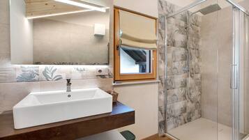 Une salle de bain avec un lavabo blanc, un miroir, une douche avec une porte en verre et une fenêtre avec un cadre en bois.