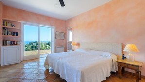 A bedroom with a large bed, a bedside table with a lamp, a bookshelf, and a view through a sliding glass door. - Villa 'Can Roca' with Sea Views, Wi-Fi and Air Conditioning (Illes Balears)