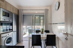 Une cuisine compacte équipée d’un four à micro-ondes, d’un four traditionnel et d’un lave-linge, une table à manger avec des chaises, et une vue à travers une porte vitrée.