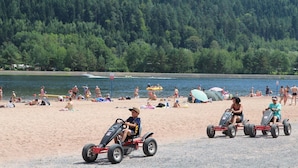 A beach with people, including children on go-karts, and a forested area in the background.