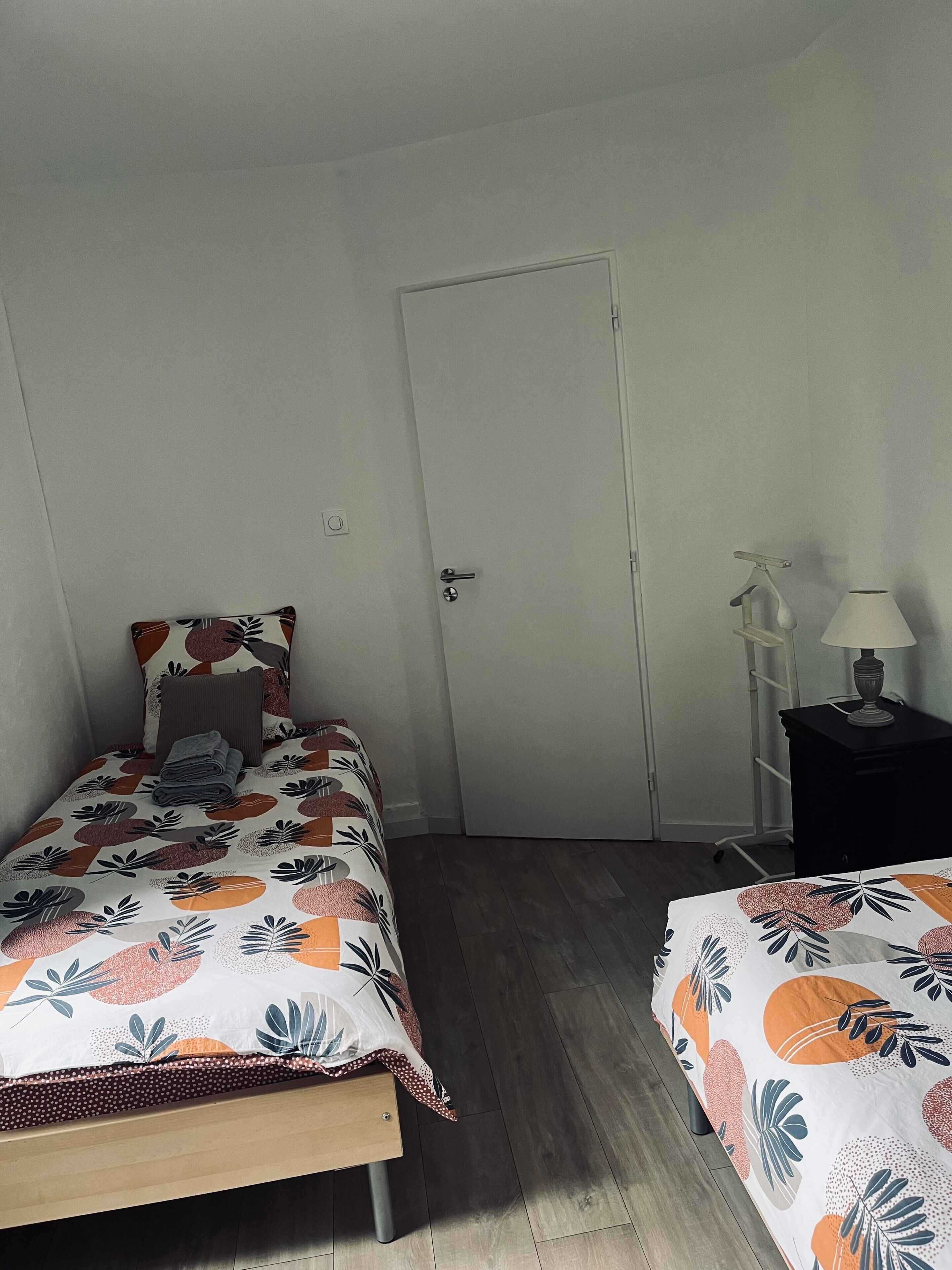 A bedroom with two beds, a door, a nightstand with a lamp, and a small white shelf.